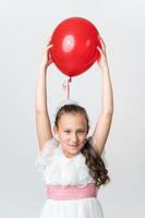 speels meisje in wit jurk houdt rood ballon bovenstaand haar hoofd met beide handen en op zoek Bij camera foto