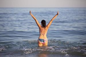 een middelbare leeftijd vrouw staat in zee water met haar armen verheven in een zwempak met een Amerikaans vlag afdrukken. foto