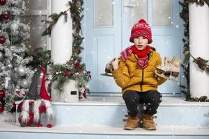 knap weinig jongen in kerstmis. grappig kind houdt oud skates en zit Aan de veranda van een huis versierd met Kerstmis boom decoraties foto