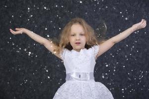 mooi weinig meisje in een wit jurk met verheven armen onder vliegend sneeuwvlokken. foto