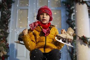 een knap jongen in helder kleren zit Aan de veranda van de huis en houdt oud schaatsen. foto