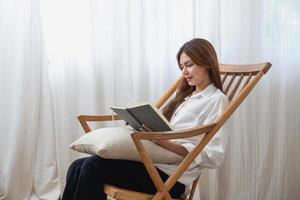 portret van een jong vrouw met een mooi gezicht vervelend een wit overhemd Holding en schrijven een dagboek Aan een notitieboekje zittend Aan een houten stoel binnen haar huis, concept van rust uit en ontspanning. foto