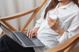 portret van een mooi jong vrouw in een wit overhemd Holding een glas van melk en een laptop naar type haar eigen verhaal en zittend Aan een houten stoel binnen de huis, de concept van rust uit foto