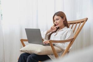 portret van een mooi jong vrouw vervelend een wit overhemd Holding een laptop naar type haar eigen verhaal en zittend Aan een houten stoel binnen de huis, de concept van rust uit en ontspanning foto