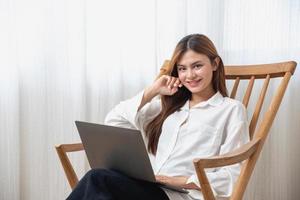 portret van een mooi jong vrouw vervelend een wit overhemd Holding een laptop naar type haar eigen verhaal en zittend Aan een houten stoel binnen de huis, de concept van rust uit en ontspanning foto