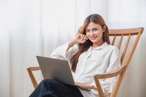 portret van een mooi jong vrouw vervelend een wit overhemd Holding een laptop naar type haar eigen verhaal en zittend Aan een houten stoel binnen de huis, de concept van rust uit en ontspanning foto