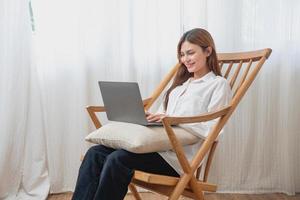 portret van een mooi jong vrouw vervelend een wit overhemd Holding een laptop naar type haar eigen verhaal en zittend Aan een houten stoel binnen de huis, de concept van rust uit en ontspanning foto