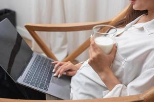 portret van een mooi jong vrouw in een wit overhemd Holding een glas van melk en een laptop naar type haar eigen verhaal en zittend Aan een houten stoel binnen de huis, de concept van rust uit foto