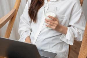 portret van een mooi jong vrouw in een wit overhemd Holding een glas van melk en een laptop naar type haar eigen verhaal en zittend Aan een houten stoel binnen de huis, de concept van rust uit foto