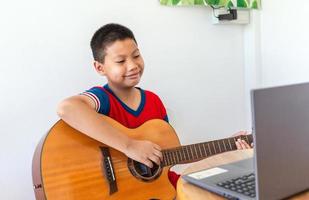 de verhaal van een jongen aan het kijken een notitieboekje computer terwijl voorbereidingen treffen naar praktijk spelen gitaar Bij huis. jongens nemen klassiek gitaar lessen online. foto