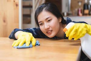 schoonmaak hygiëne, glimlach Aziatisch jong hulp in de huishouding, serveerster vrouw vervelend geel beschermend handschoenen, schoonmaak Aan hout tafel, gebruik blauw vod afvegen naar stof en verstuiven in restaurant. huishouding schoonmaken, schoner. foto