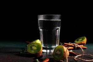 een glas van verkoudheid water Aan een groen achtergrond. versierd met plak kiwi, kaneel stokken, badyan, munt bladeren. kopiëren ruimte foto