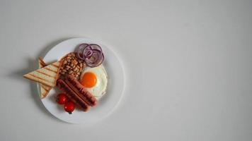 top visie Engels ontbijt. gebakken ei, twee gebakken worstjes, gegrild brood geroosterd brood, ingeblikt bonen, blauw uien en kers tomaten Aan een wit achtergrond foto