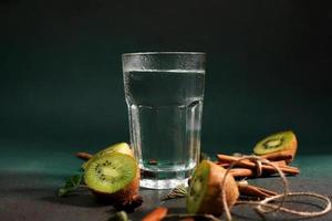 een glas van verkoudheid water Aan een groen achtergrond. versierd met plak kiwi, kaneel stokken, badyan, munt bladeren. kopiëren ruimte foto