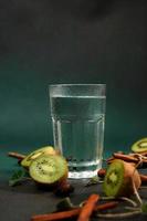 een glas van verkoudheid water Aan een groen achtergrond. versierd met plak kiwi, kaneel stokken, badyan, munt bladeren. kopiëren ruimte foto