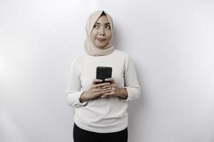 een attent jong Aziatisch moslim vrouw vervelend een hijab en Holding haar kin terwijl pratend Aan de telefoon geïsoleerd door een wit achtergrond foto