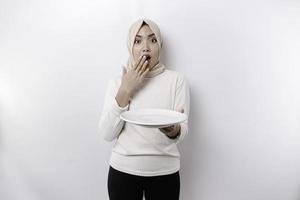 een geschokt Aziatisch moslim vrouw is vastend en hongerig en Holding een bord met haar mond breed Open foto