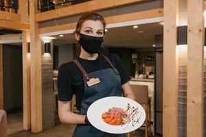 een stijlvolle serveerster die een zwart gezichtsmasker en wegwerphandschoenen draagt, houdt een salade met munt en dolendwitz vast in een restaurant foto