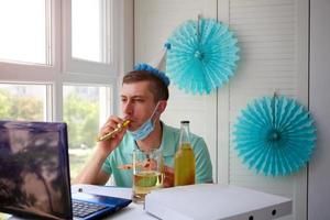 een Mens in een medisch masker zit in voorkant van een laptop en slagen fluitjes, viert verjaardag met vrienden over- de internetten. foto