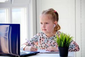 meisje schoolmeisje in hoofdtelefoons looks Bij een laptop en schrijft in een notebook. afstand aan het leren, studie Bij huis, online. foto