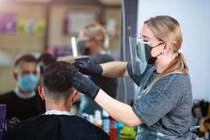 een kapper met veiligheid maatregelen voor covid19, bezuinigingen haar- met een schaar naar een Mens in een geneeskunde masker, sociaal afstand, snijdend haar- met een medisch masker, gezicht schild , handschoenen foto