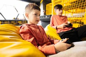 twee broers spelen video spel troosten, zittend Aan geel poef in kinderen Speel centrum. foto