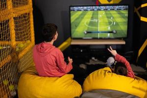 twee broers spelen Amerikaans voetbal video spel troosten, zittend Aan geel poef in kinderen Speel centrum. foto