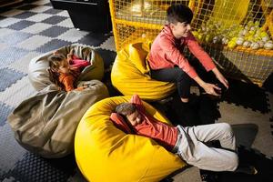 drie kinderen spelen video spel troosten, zittend Aan geel poef in kinderen Speel centrum. foto