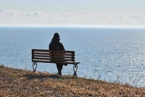 single meisje in een zwart jasje en hoed zittend Aan bank Bij klif Bij voorkant van zee vredig stil plaats foto