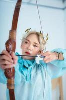 een Aziatisch vrouw met blond haar- Holding een pijl terwijl vervelend een blauw jurk foto