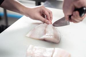 gefocust mannetje chef in uniform snijdend vis filet Aan bord, chef bezuinigingen wit tilapia vis in keuken. foto