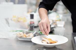 gefocust hand- mannetje chef in zwart uniform versieren voedsel, bestrooi garnituren Aan Koken gebakken steak vis filet , wit tilapia in wit bord in keuken. foto