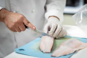 gefocust mannetje chef in uniform snijdend vis filet Aan bord, chef bezuinigingen wit tilapia vis in keuken. foto