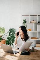 bedrijf Aziatisch vrouw pratend Aan de telefoon en gebruik makend van een laptop met een glimlach terwijl zittend Bij huis kantoor foto