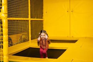 weinig meisje kind tonen duim omhoog Aan trampoline Bij geel speelplaats park. kind in actief vermaak. foto