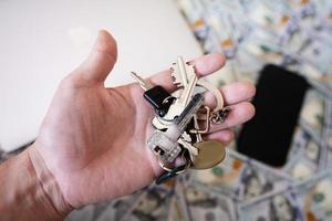 Mens hand- houden sleutels Aan dollar geld, laptop en telefoon achtergrond. foto