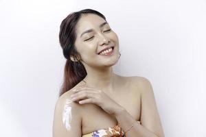 huid zorg producten concept. Aziatisch vrouw toepassen hydraterend lotion Aan lichaam na douche, staand verpakt in handdoek, bijgesneden beeld foto