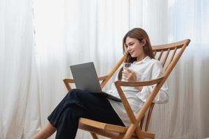 portret van een mooi jong vrouw in een wit overhemd Holding een glas van melk en een laptop naar type haar eigen verhaal en zittend Aan een houten stoel binnen de huis, de concept van rust uit foto