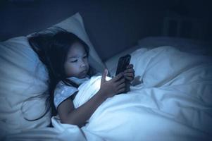 Aziatisch kind spelen spel Aan smartphone in de bed Bij nacht, de meisje verslaafde sociaal media foto