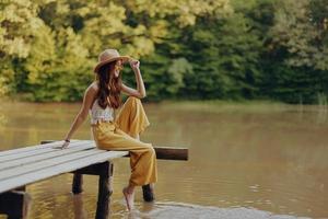 een jong gelukkig hippie vrouw zittend Aan de meer kust Aan een brug in eco kleding gemaakt van natuurlijk materialen in harmonie met natuur in de vallen foto
