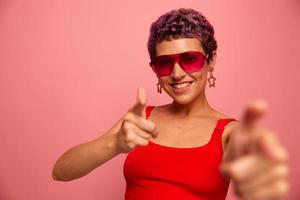 mode portret van een vrouw met een kort kapsel in gekleurde zonnebril met ongebruikelijk accessoires met oorbellen glimlacht Aan een roze helder achtergrond shows haar handen naar de camera foto