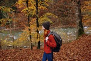 kant visie van gelukkig vrouw in park in herfst in de buurt rivier- en rugzak Aan haar terug foto