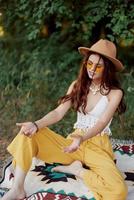 een jong hippie vrouw in eco-kleding heeft pret lachend en ineenkrimpen zittend Aan een plaid buiten in de vallen met een hoed en geel zonnebril. reizen levensstijl foto