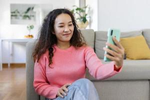 aantrekkelijk glimlachen Aziatisch vrouw gebruik makend van slim telefoon terwijl zittend Aan de sofa Bij huis. communicatie en video telefoontje concept. foto