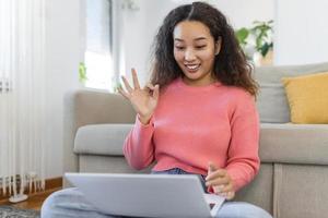 gelukkig jong Aziatisch vrouw hebben pret aan het doen video telefoontje gebruik makend van laptop in haar huis, golvend hand- video conferentie roeping Aan laptop computer zitten Aan sofa afstand leren zoom online virtueel vergadering Bij huis. foto