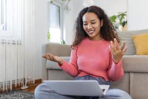 gelukkig jong Aziatisch vrouw hebben pret aan het doen video telefoontje gebruik makend van laptop in haar huis, golvend hand- video conferentie roeping Aan laptop computer zitten Aan sofa afstand leren zoom online virtueel vergadering Bij huis. foto