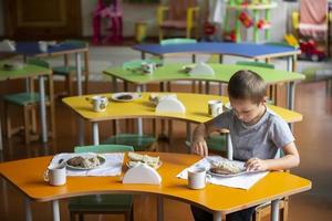 een kind eet in kleuterschool. een weinig jongen Bij de tafel met voedsel. ontbijt voor kinderen. foto