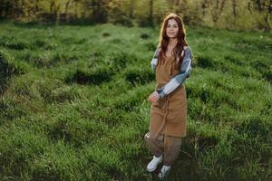 een vrouw tuinman in een schort staat in een veld- van groen gras buitenshuis, glimlachen Aan een zomer middag in een zonnig zonsondergang na een dag werk foto