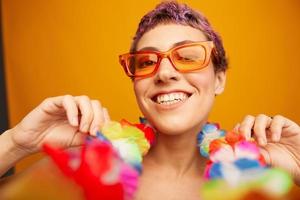 portret van een vrouw met een bloemen hawaiiaans krans in de omgeving van haar nek is hebben pret dansen en glimlachen in helder kleren Aan een oranje achtergrond vervelend zonnebril, levensstijl partij hawaiiaans stijl foto