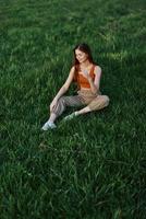 freelancer vrouw genieten van de buitenshuis zittend in de park Aan de groen gras in gewoontjes kleding met lang rood haar- lit door de helder zomer zon zonder muggen foto